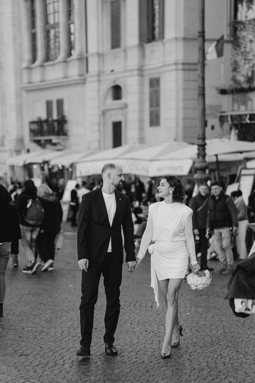 Black and white photo of a stylish couple holding hands walking through a busy European plaza, bride in short white dress.