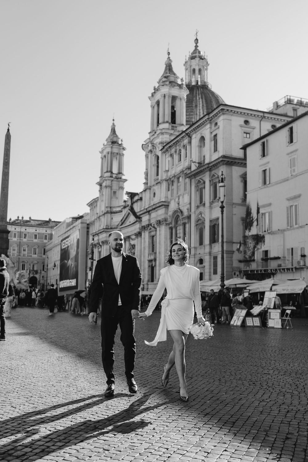 Newlyweds walk hand in hand across cobblestone Piazza Navona in Rome with historic baroque church backdrop.