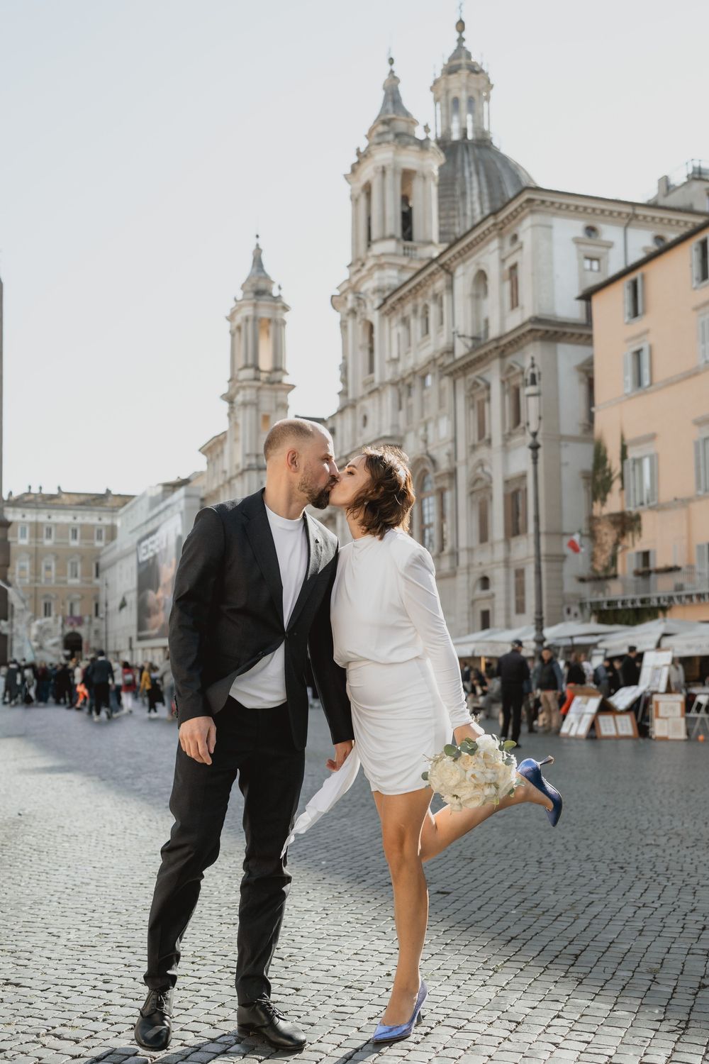 Couple sharing a romantic kiss in Piazza Navona, Rome, bride in white dress holding bouquet, groom in black suit.