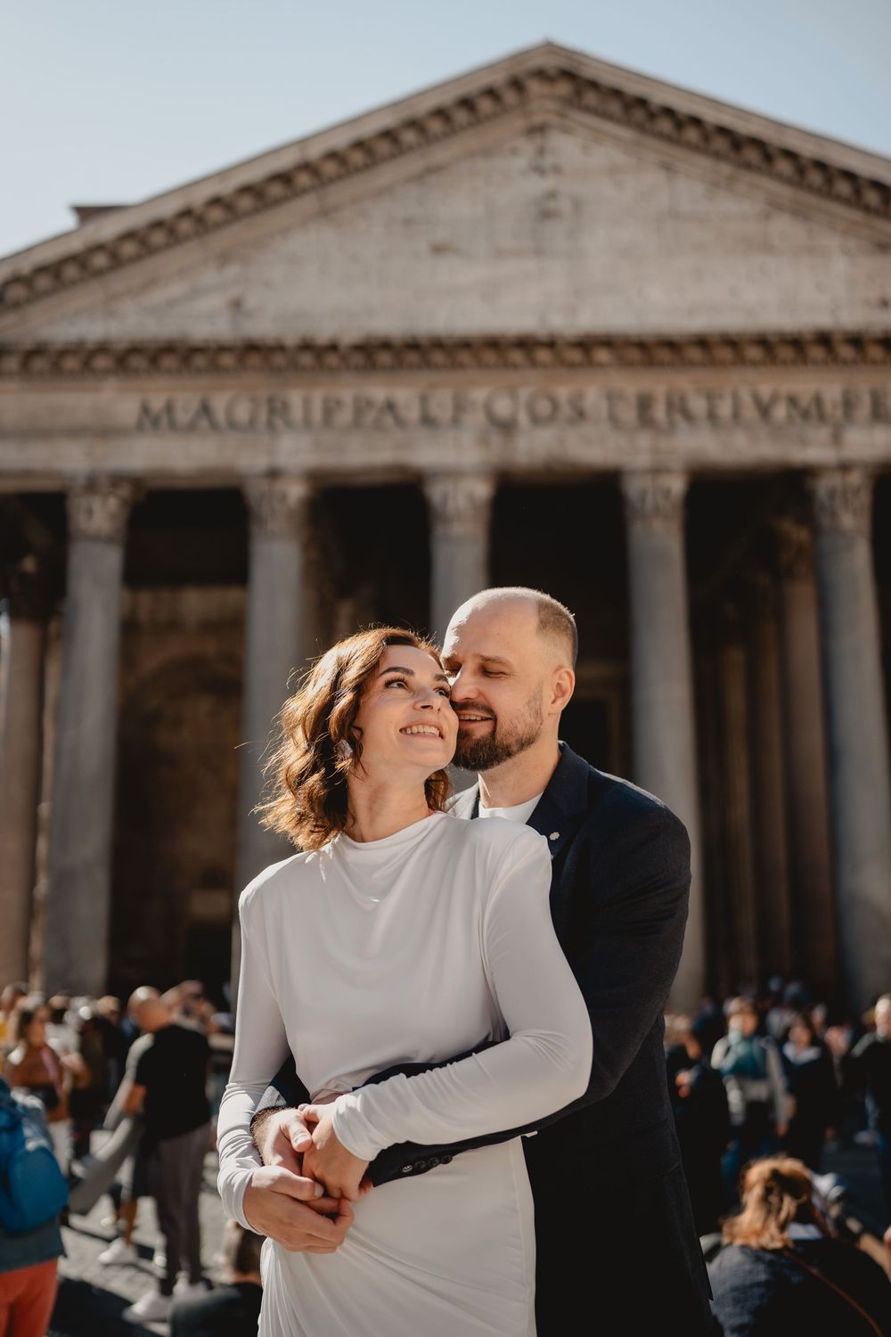 Couple embracing romantically in front of the Pantheon in Rome, Italy, with ancient columns and Latin inscription visible.