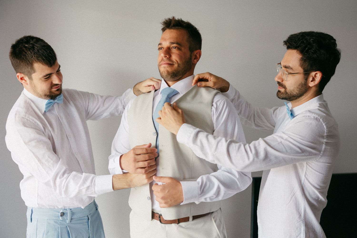 Two groomsmen help the groom adjust his tie and suit jacket while preparing for a wedding ceremony.