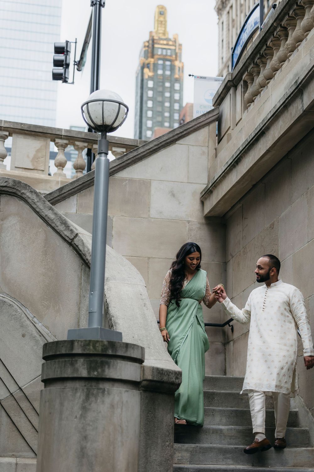 Couple in traditional Indian attire holding hands during engagement session on stone steps with Chicago skyline in background.