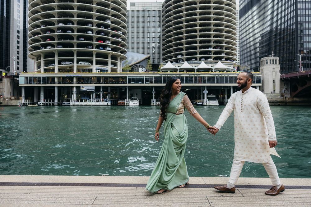 Indian couple in traditional attire holding hands during engagement session by Chicago River with Marina City towers in background.