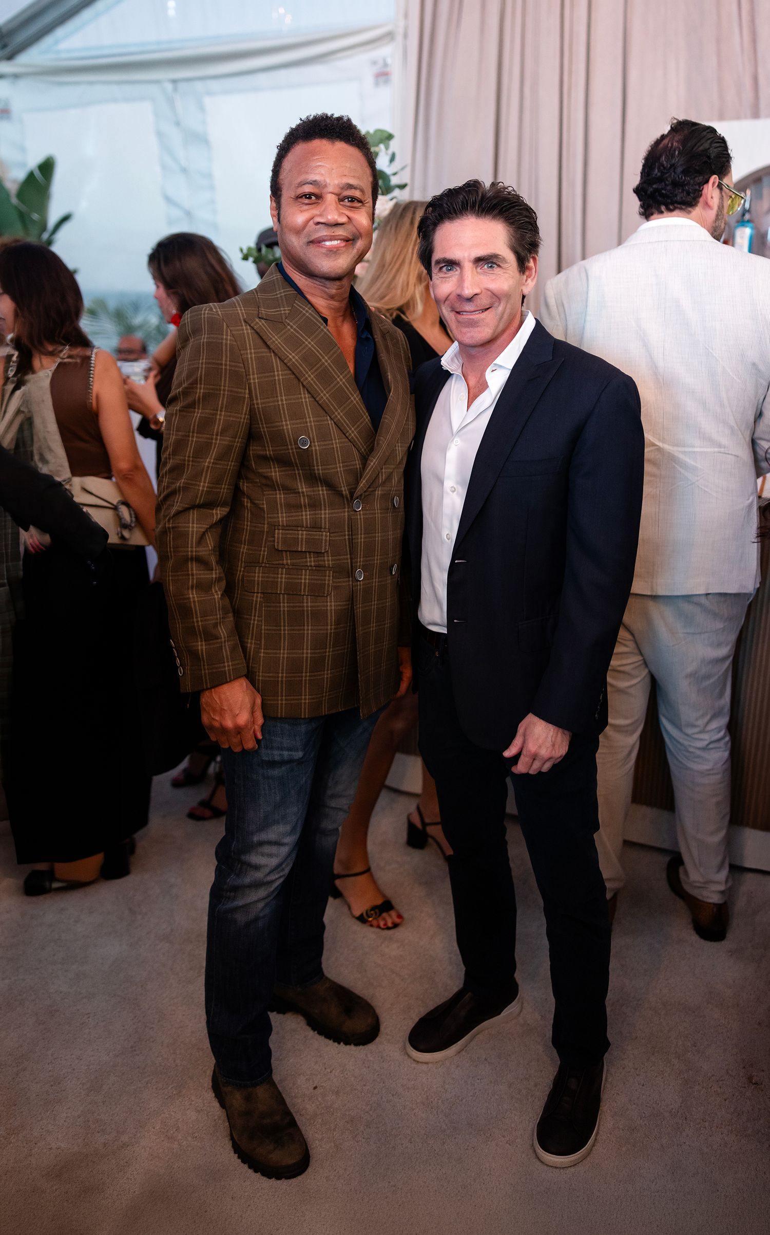 Two men in blazers smile and pose together at an upscale indoor social event with guests in background.