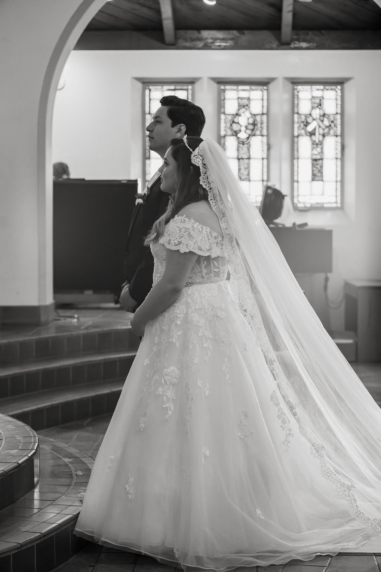 Black and white photo of a bride in lace gown and veil with groom in church with stained glass windows.