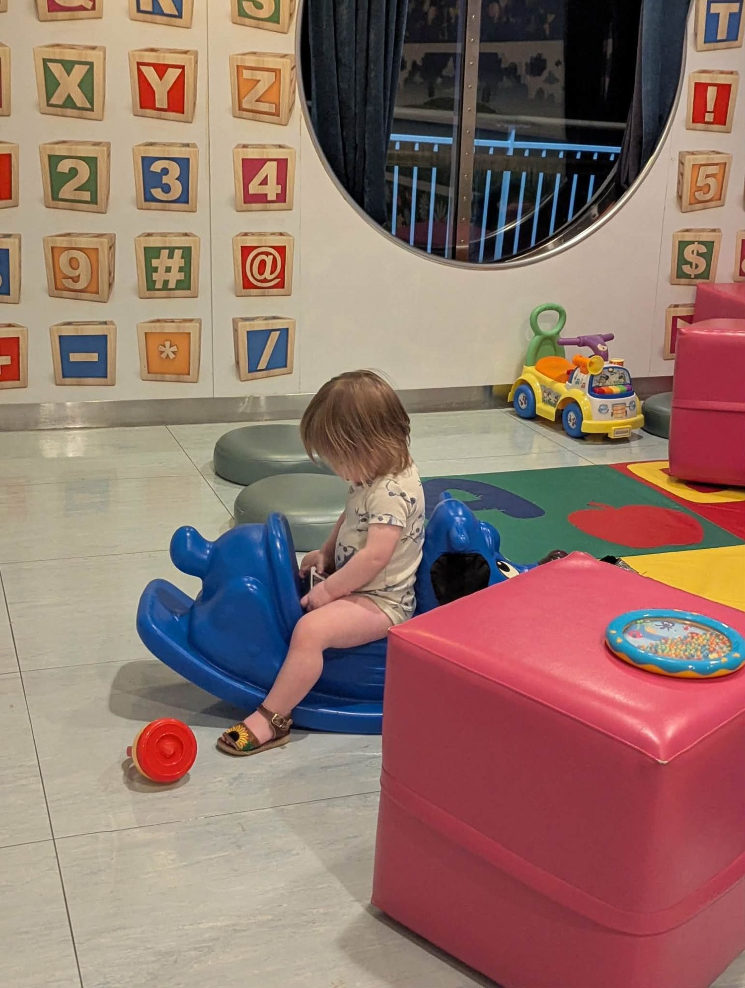 Toddler playing on a blue rocking toy in a colorful children's play area with alphabet block wall decor.