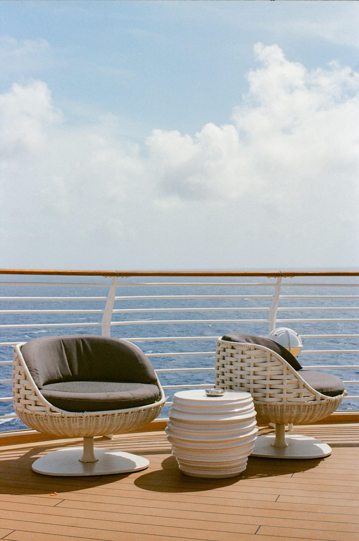 Modern wicker chairs and striped side table on a cruise ship deck overlooking the ocean under a partly cloudy sky.