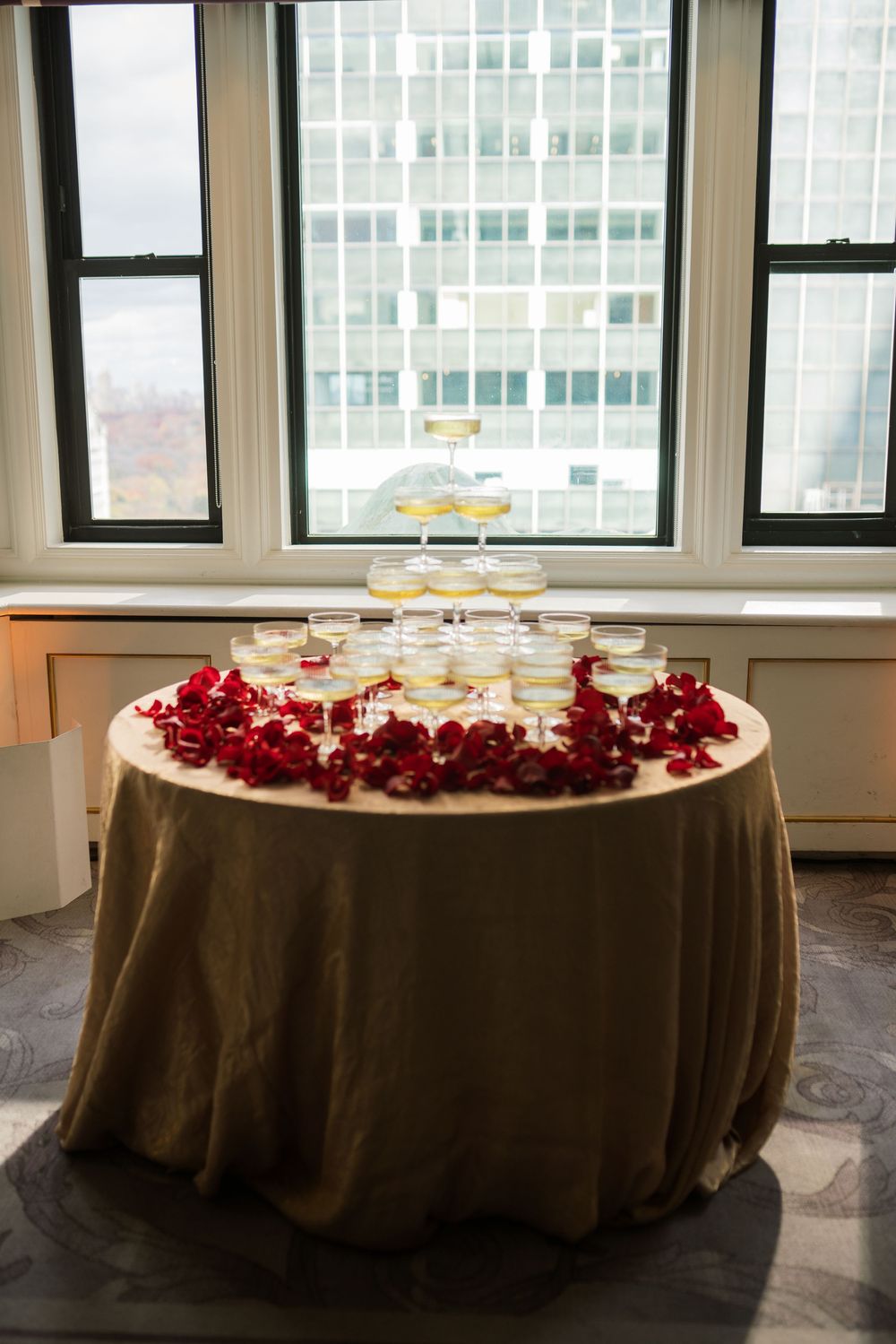 Elegant champagne tower with red rose petals on a gold tablecloth, set near large windows with city views.