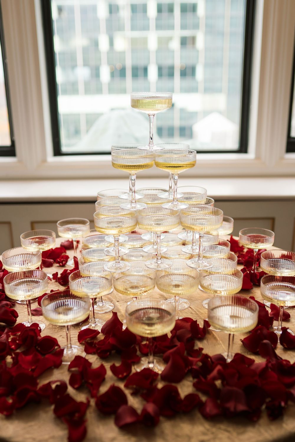 Pyramid champagne tower with coupe glasses surrounded by red rose petals on a marble table at a wedding.