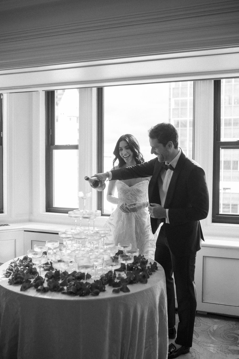 Black and white photo of bride and groom pouring champagne tower at elegant wedding reception.