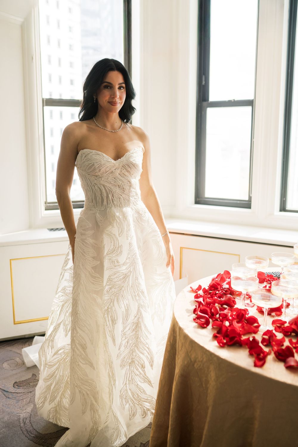 Bride in strapless white lace wedding gown standing near window beside table with red rose petals.