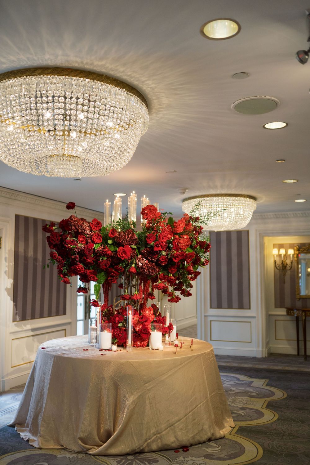 Elegant wedding sweetheart table with lush red floral centerpiece under crystal chandeliers in a luxury ballroom.