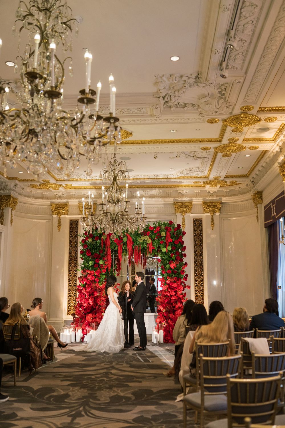 Elegant wedding ceremony in ornate ballroom with red floral arch, crystal chandeliers, and guests seated in gold chairs.