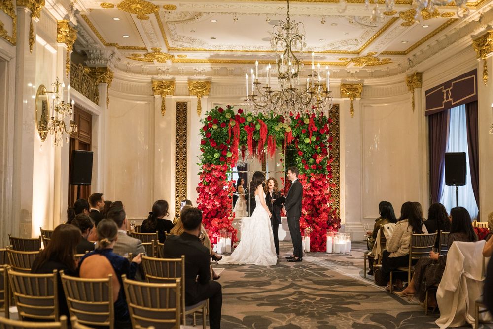 Elegant wedding ceremony in ornate ballroom with red Christmas trees, floral arch, chandelier, and guests seated in gold chairs.