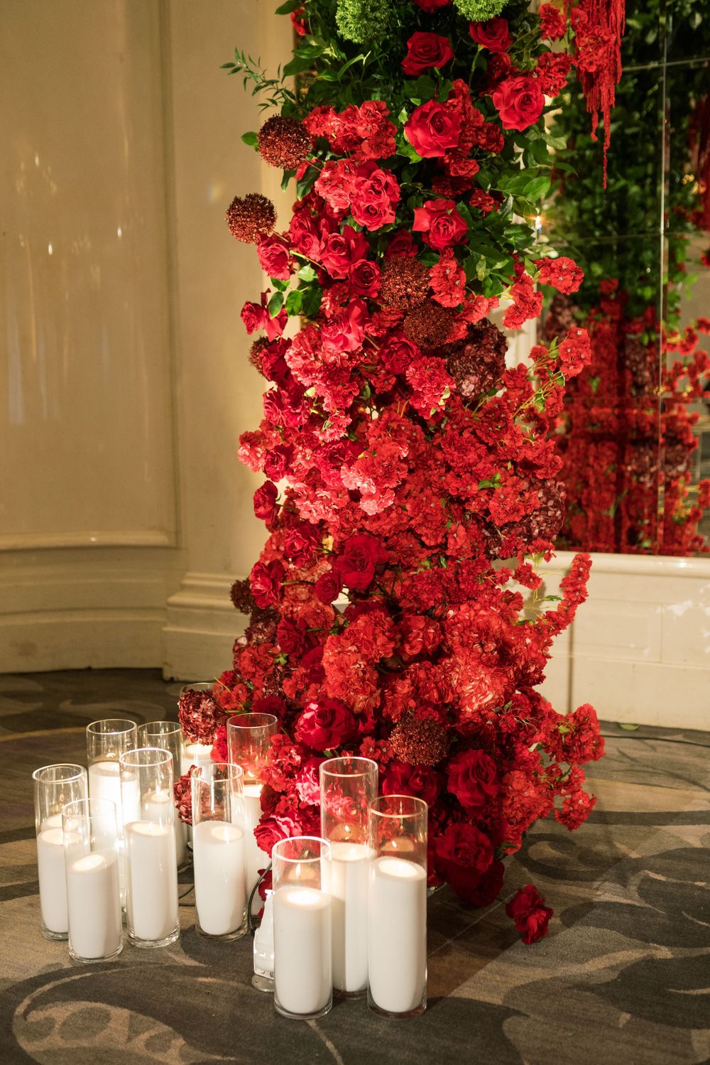 Lush red floral wall display with roses and carnations, accented by glowing white pillar candles on a patterned floor.