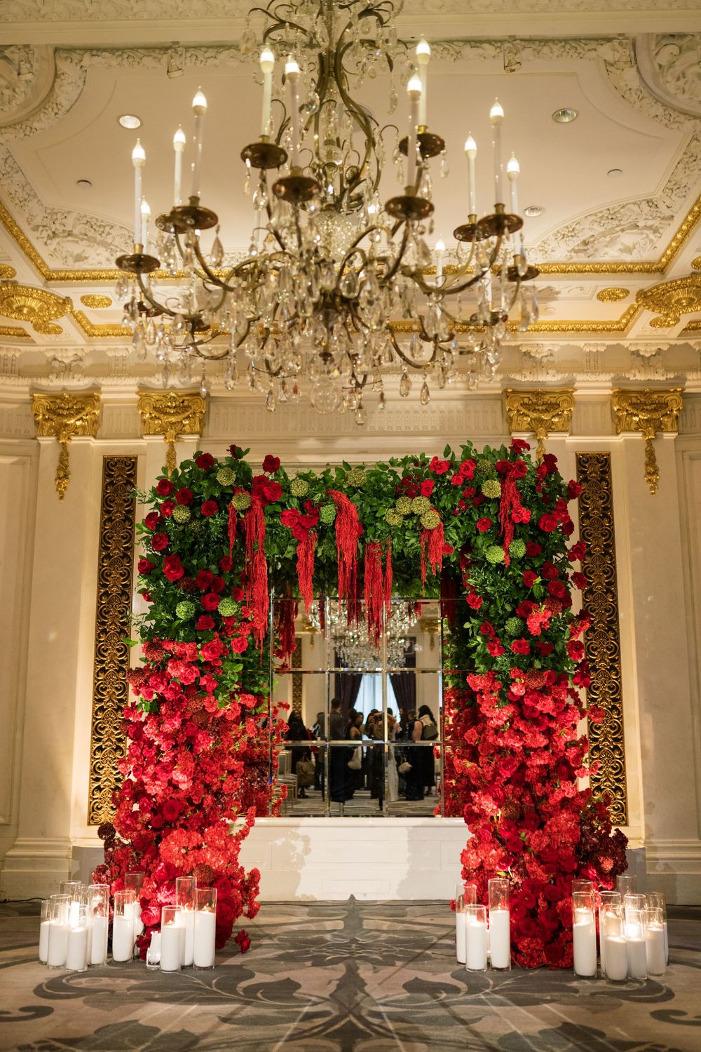 Elegant floral arch with red roses and greenery framing a window, flanked by candles under a crystal chandelier.