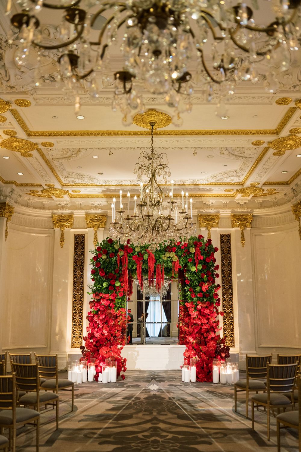 Elegant wedding ceremony venue with red floral arch, crystal chandeliers, and candlelit aisle in a grand ballroom.