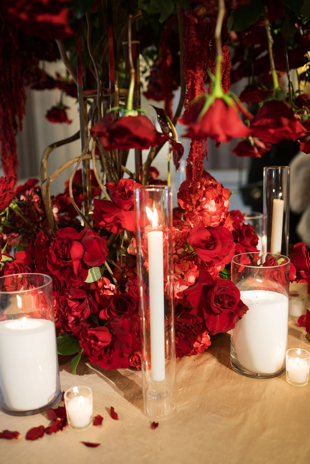 Elegant wedding centerpiece with red roses, carnations, candles in glass holders, and hanging floral arrangements on a draped table.