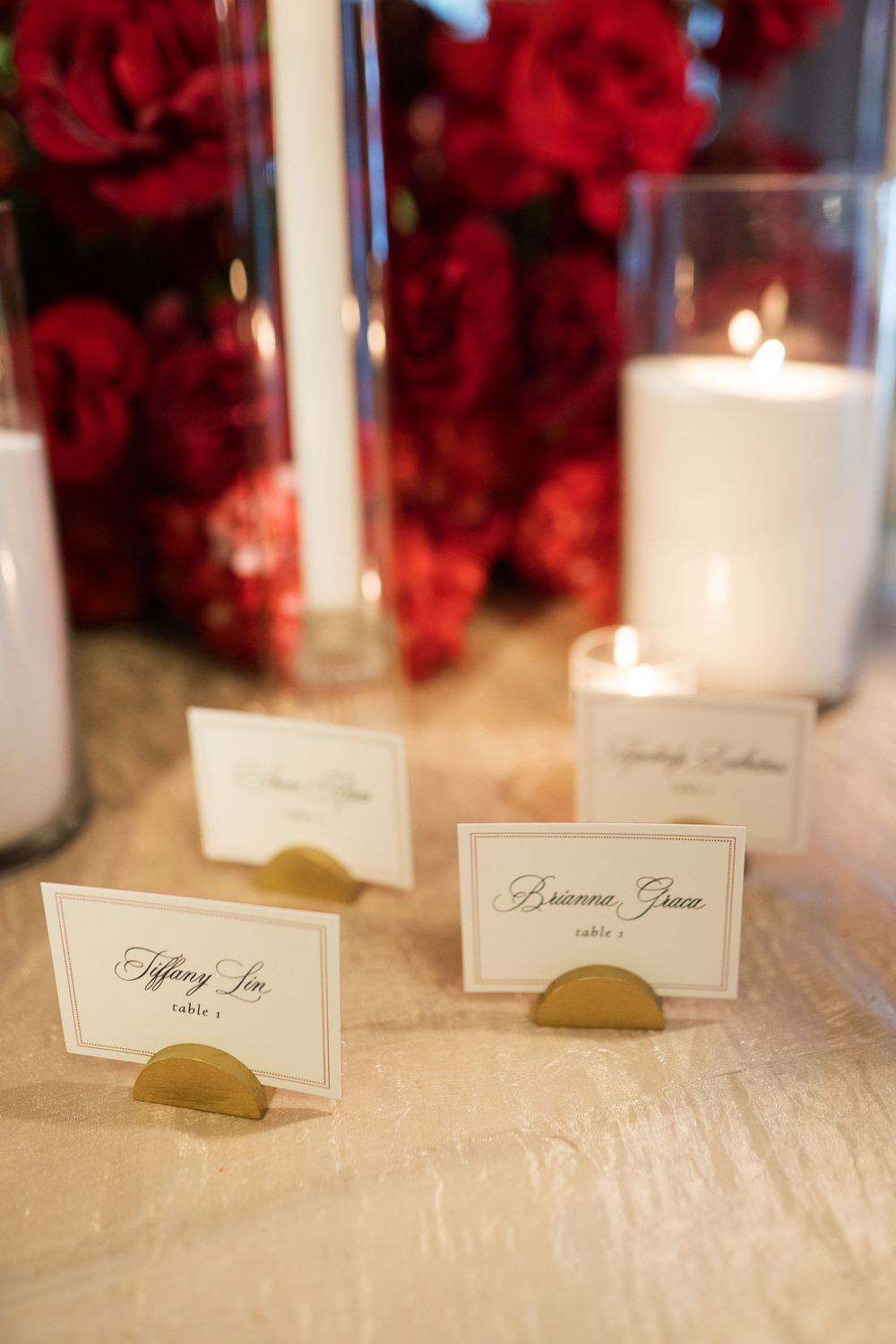Gold place card holders with calligraphy escort cards surrounded by red roses and glowing candles at wedding reception.