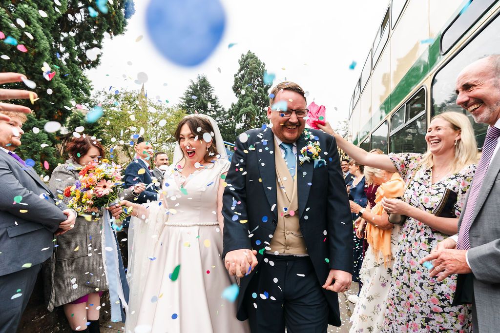 Bride and groom walk through colorful confetti shower surrounded by cheering wedding guests outdoors.