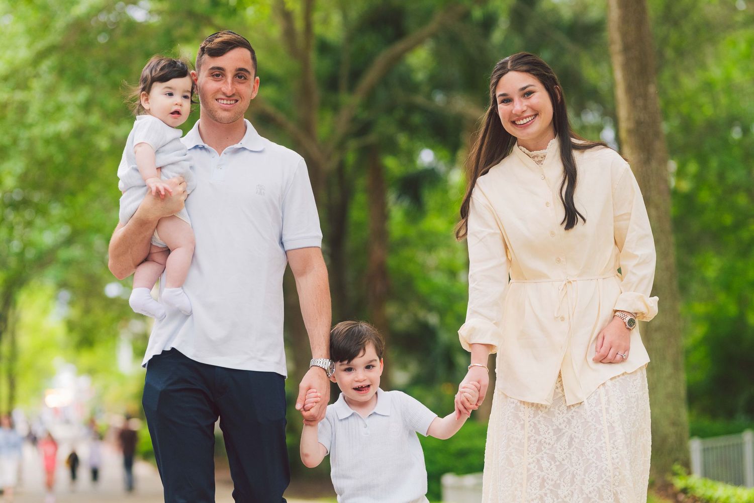Happy family of four walking outdoors in a lush green park, parents holding children's hands on a sunny day.