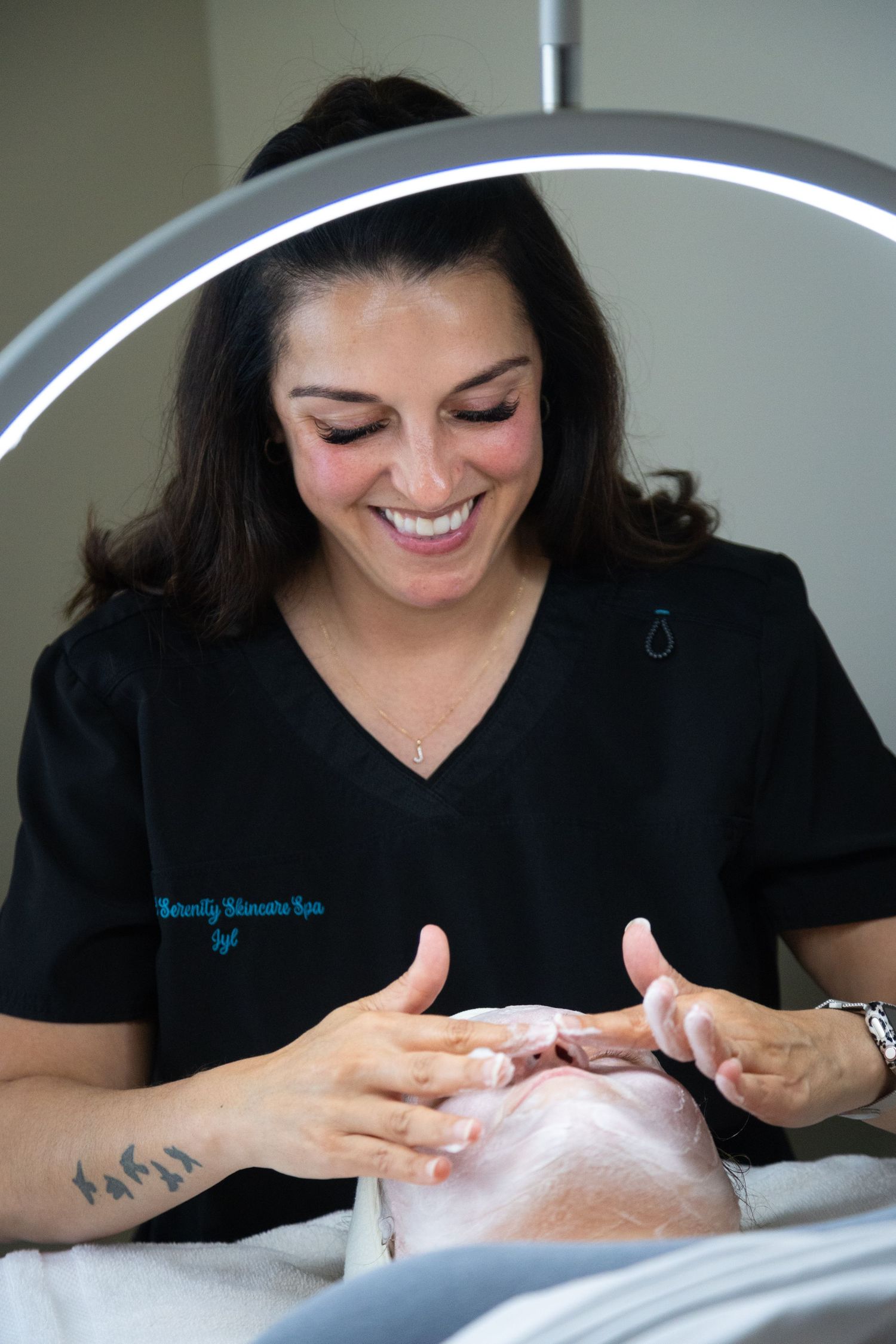 Smiling esthetician in black scrubs performing a facial treatment on a client at Naturally Skincare Spa.