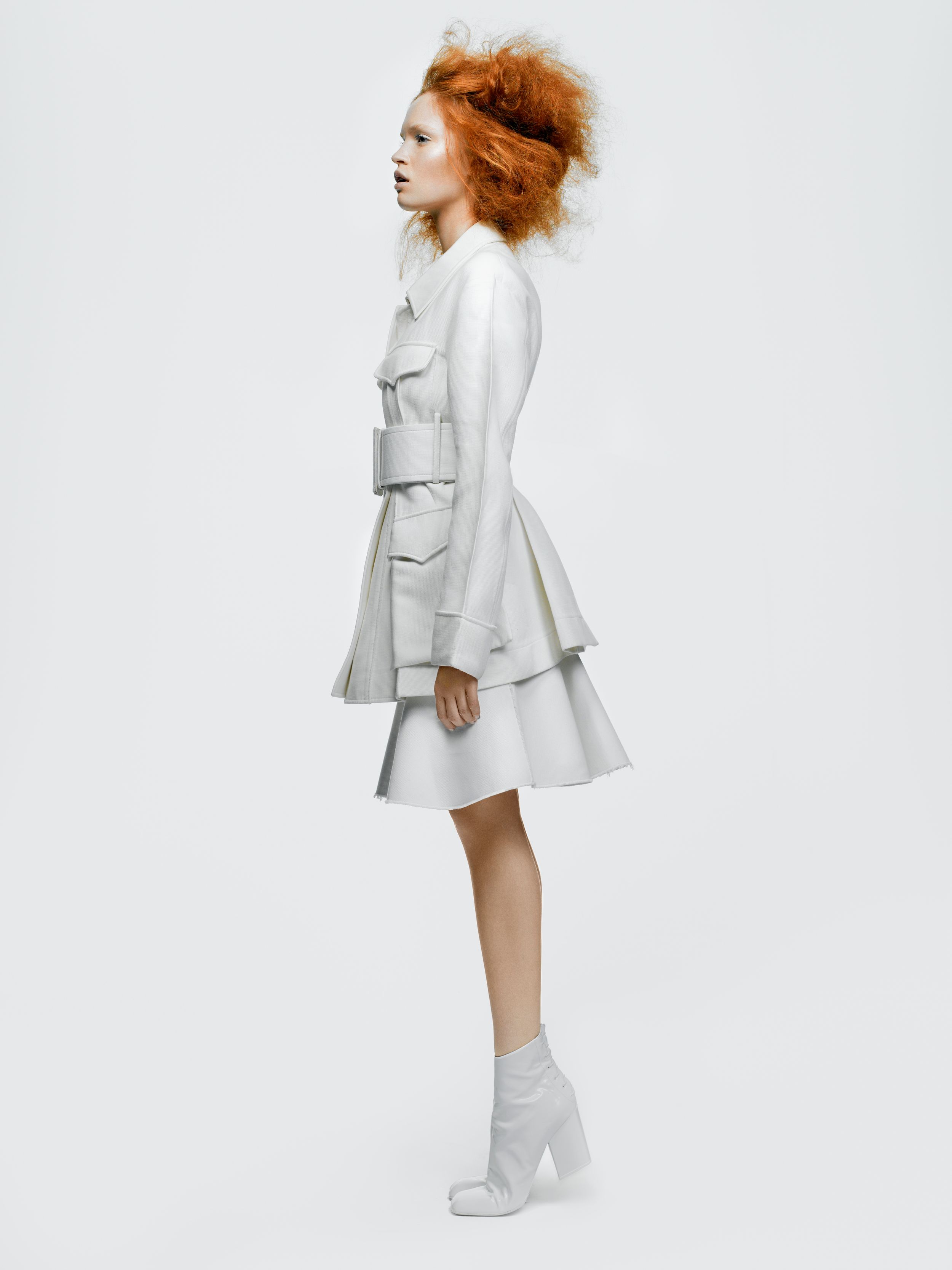 Woman in white tiered ruffle dress and ankle boots with red curly hair poses against white background.