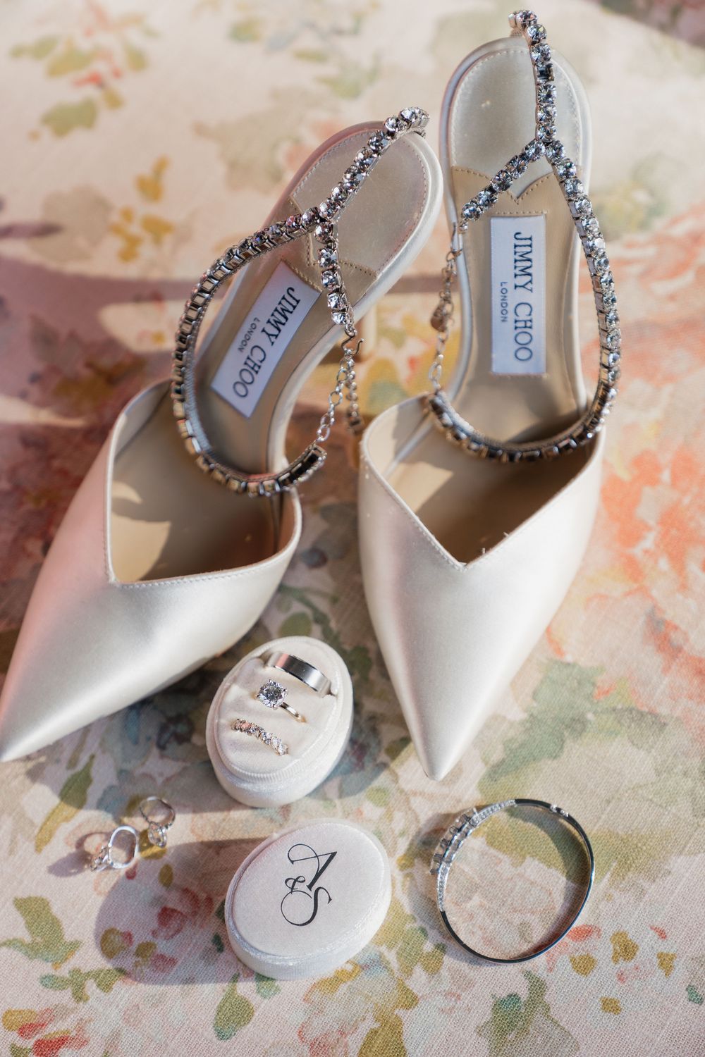 White Jimmy Choo bridal heels with crystal straps displayed with wedding rings and accessories on floral fabric.