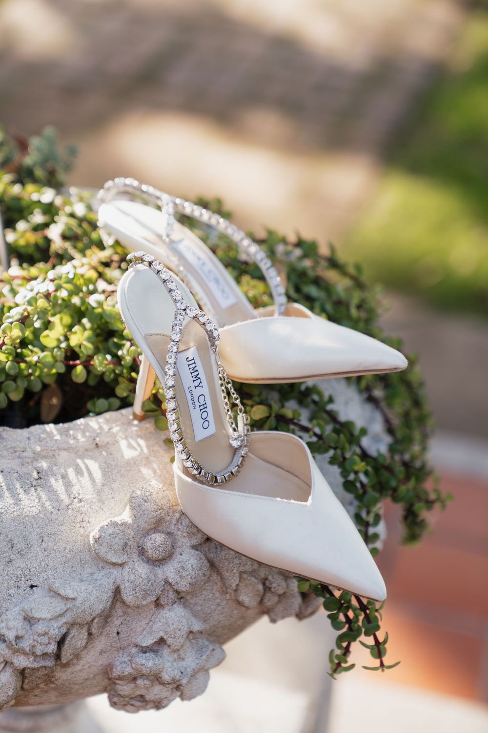 White Jimmy Choo crystal-strap bridal heels displayed on a stone planter with green foliage backdrop.