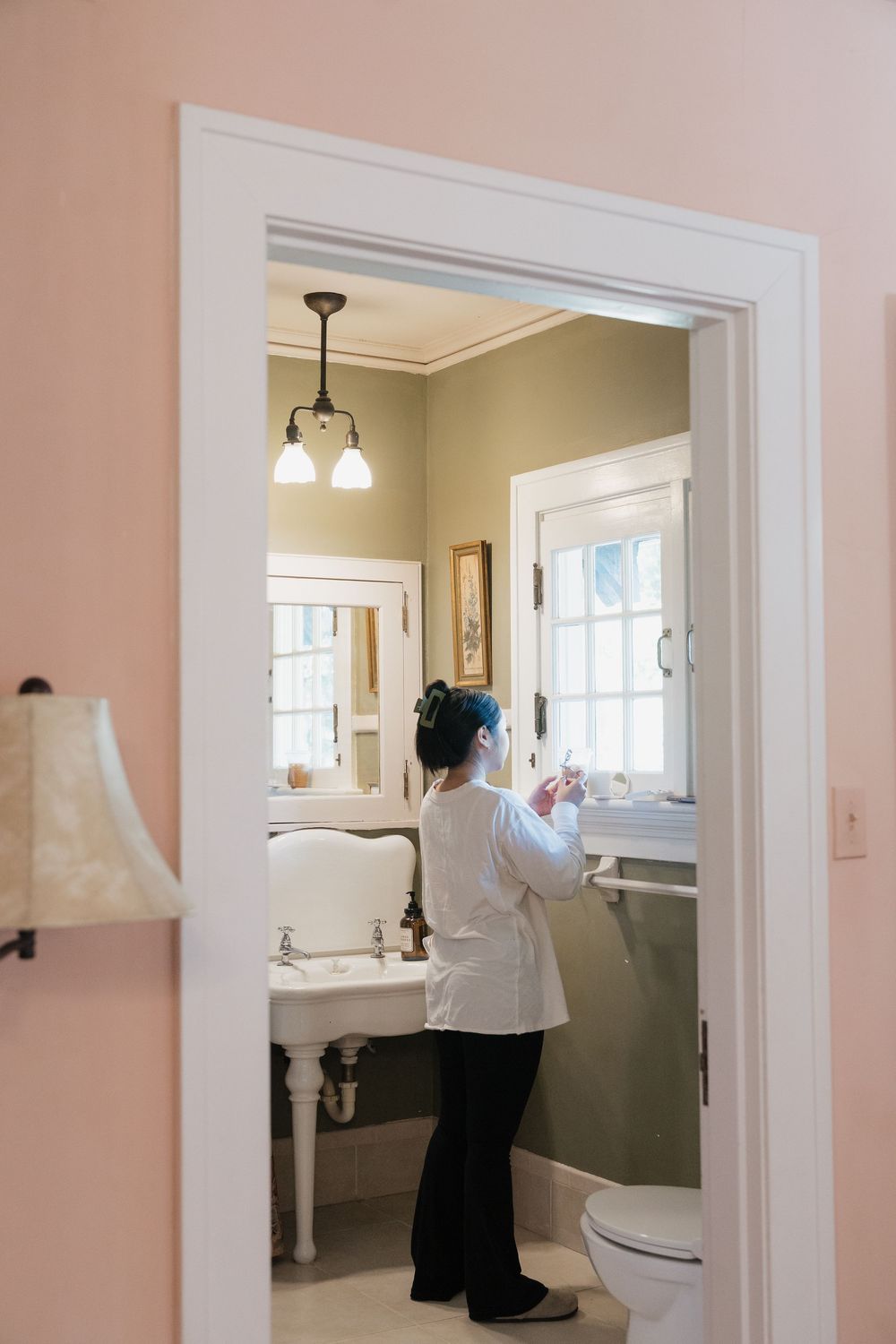 Woman in white coat stands at vintage pedestal sink in elegant pink and sage green bathroom viewed through doorway.