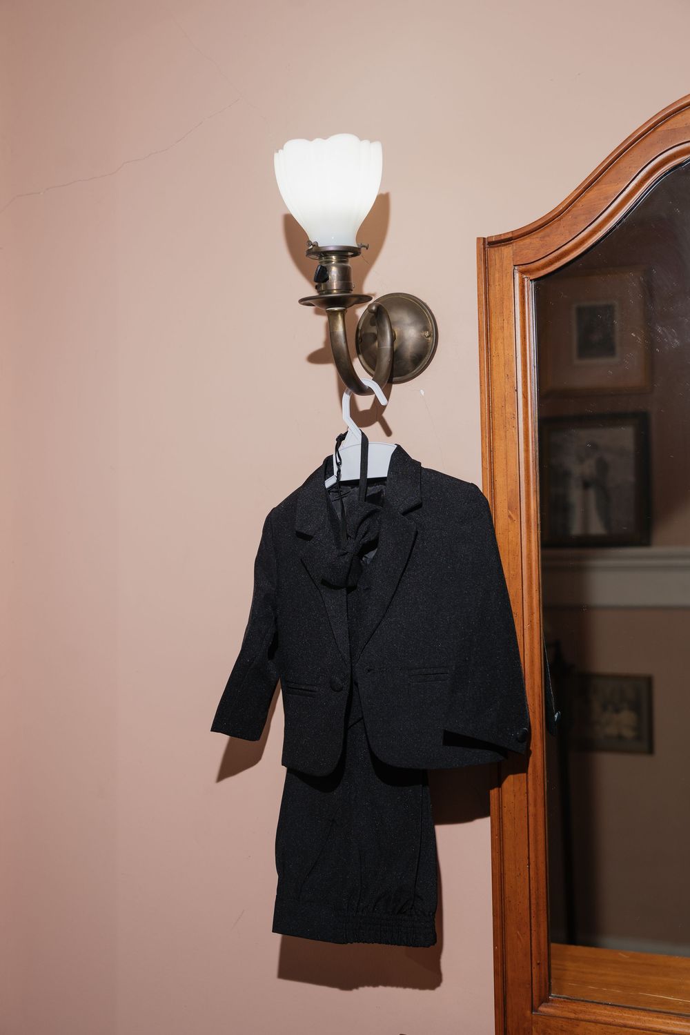 Small black child's tuxedo suit hanging on ornate brass wall sconce beside wooden framed mirror.