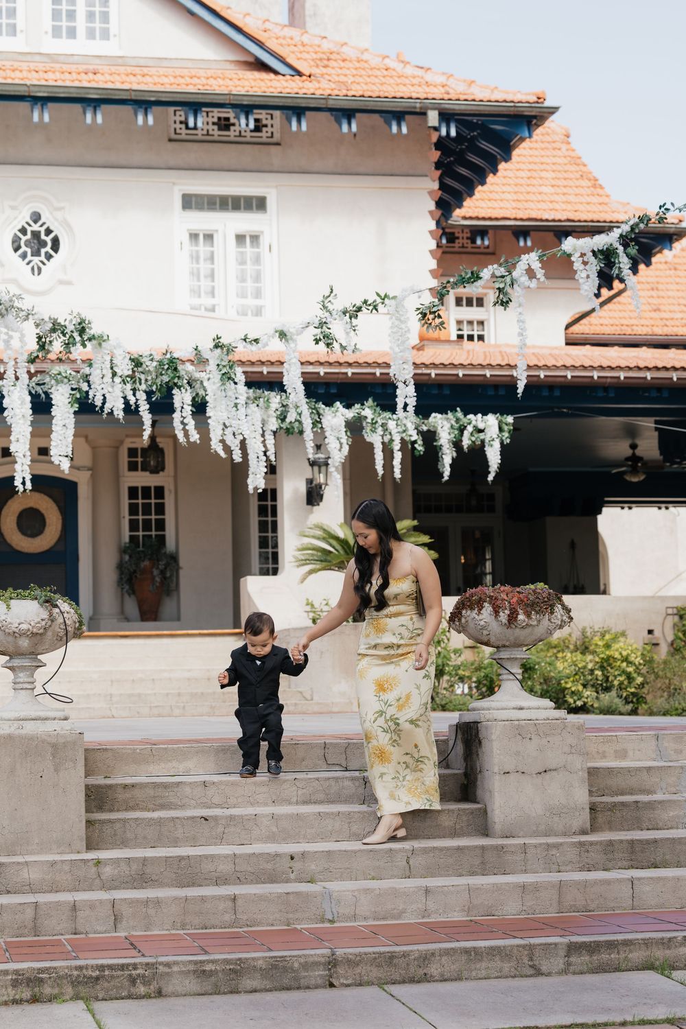 Woman in floral dress holds toddler's hand on stone steps outside white villa with hanging wisteria decorations.