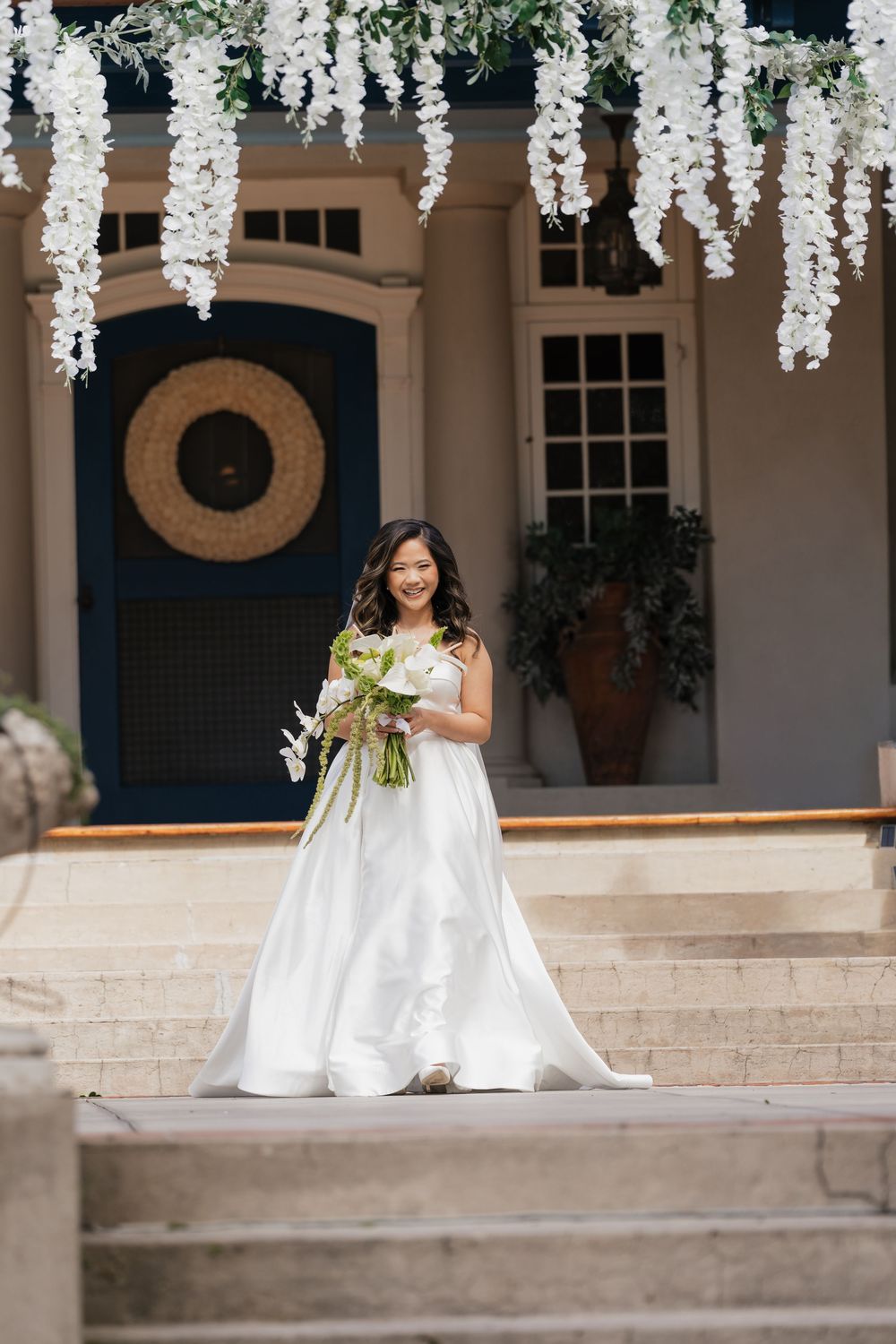 Smiling bride in white ball gown holds floral bouquet on steps beneath hanging white wisteria at elegant venue.