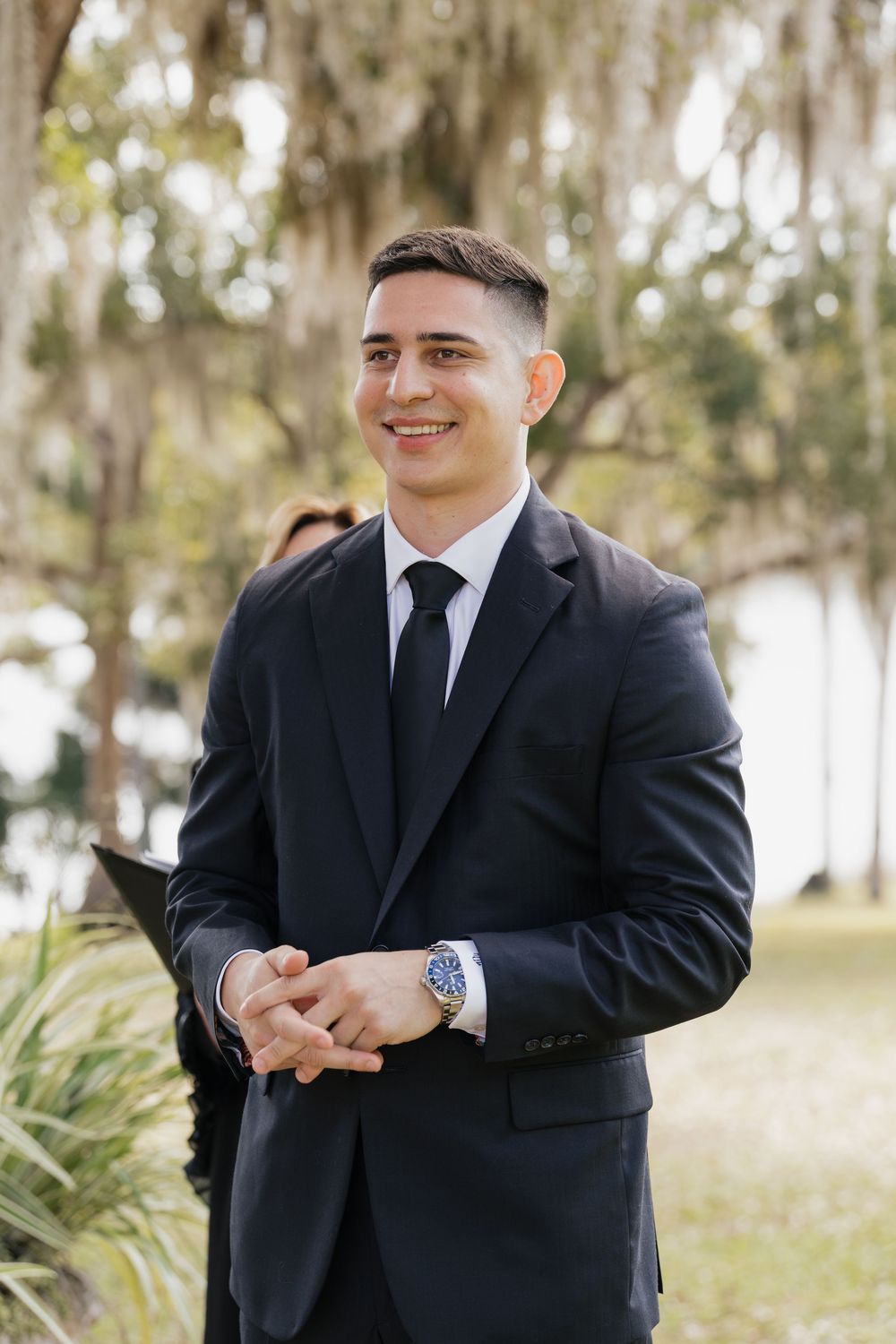 Smiling groom in dark suit and black tie clasping hands outdoors at wedding ceremony.