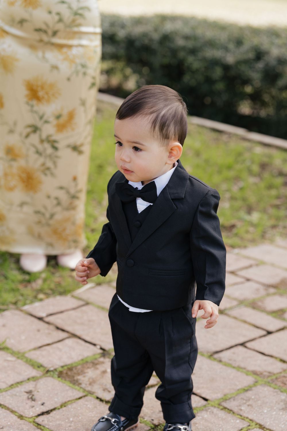 Adorable baby boy in black tuxedo with bow tie standing on stone pathway at outdoor wedding.