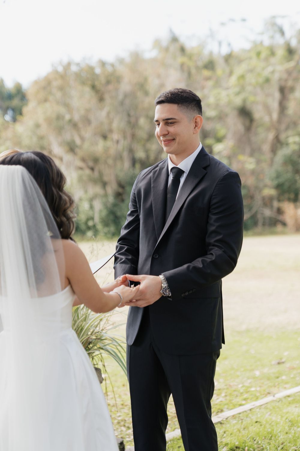 Groom in black suit smiles warmly while holding brides hands during outdoor wedding ceremony.