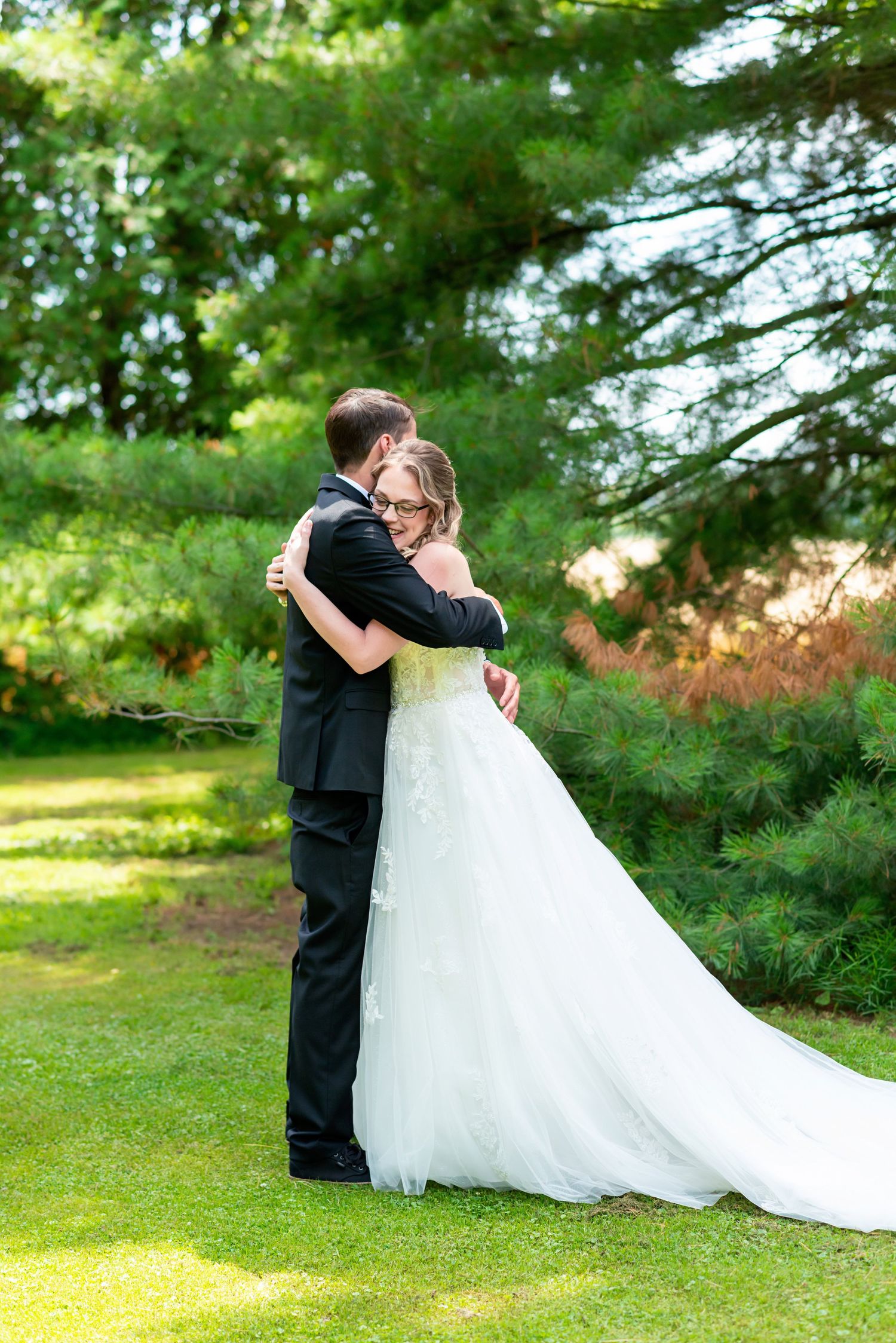 Bride and groom embrace in lush green garden, bride in white gown with train, groom in black suit.