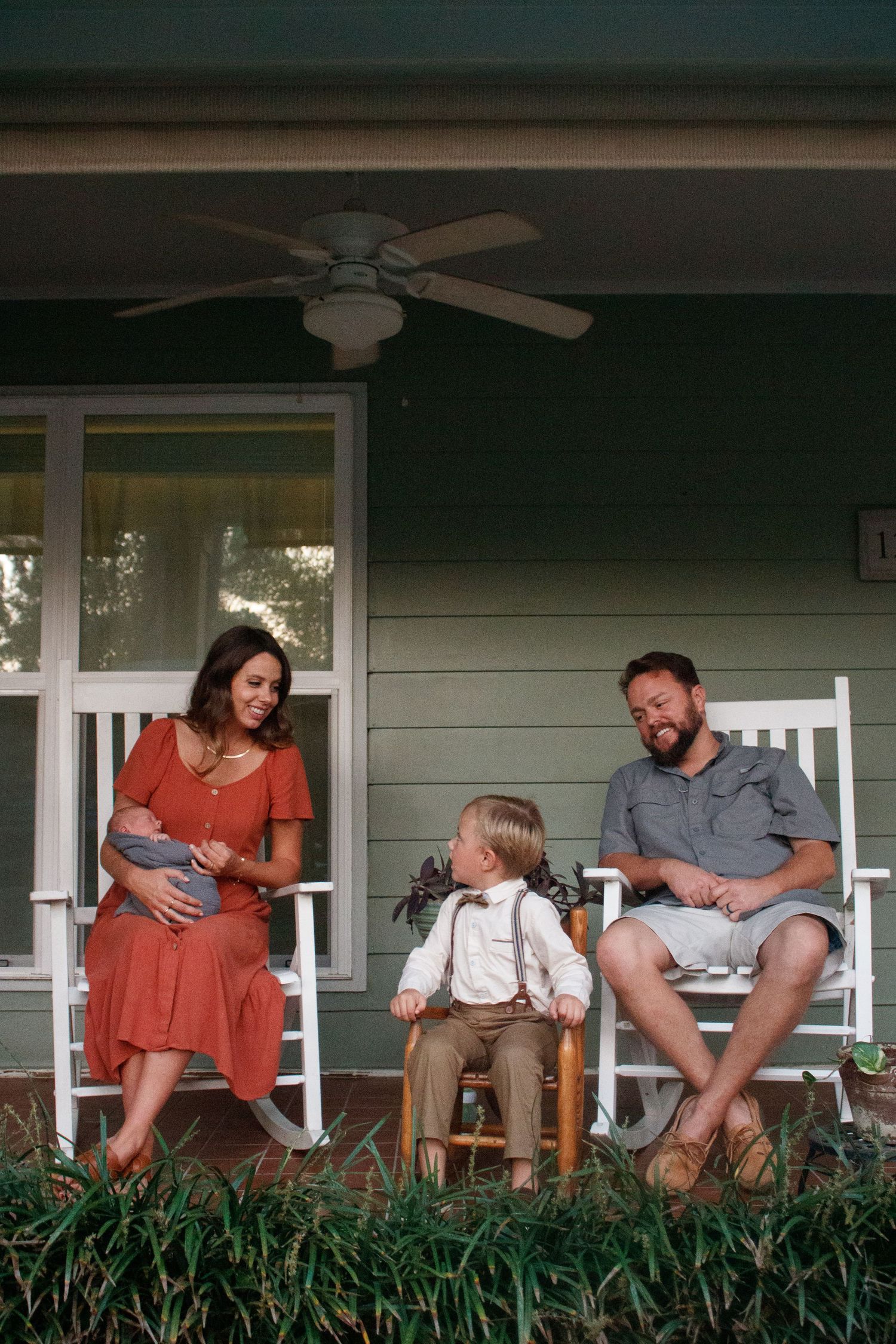 Family of four relaxing on a green porch with parents holding a newborn and a toddler in a bow tie.