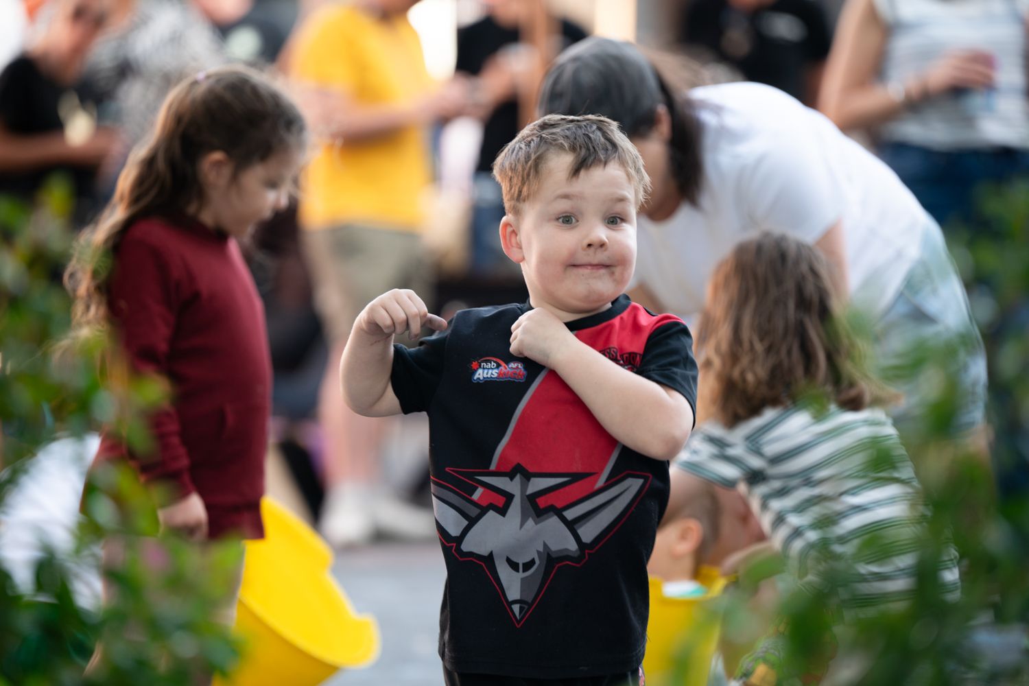 Smiling young boy in a sports jersey flexes his arm at a fun outdoor community event with yellow buckets.