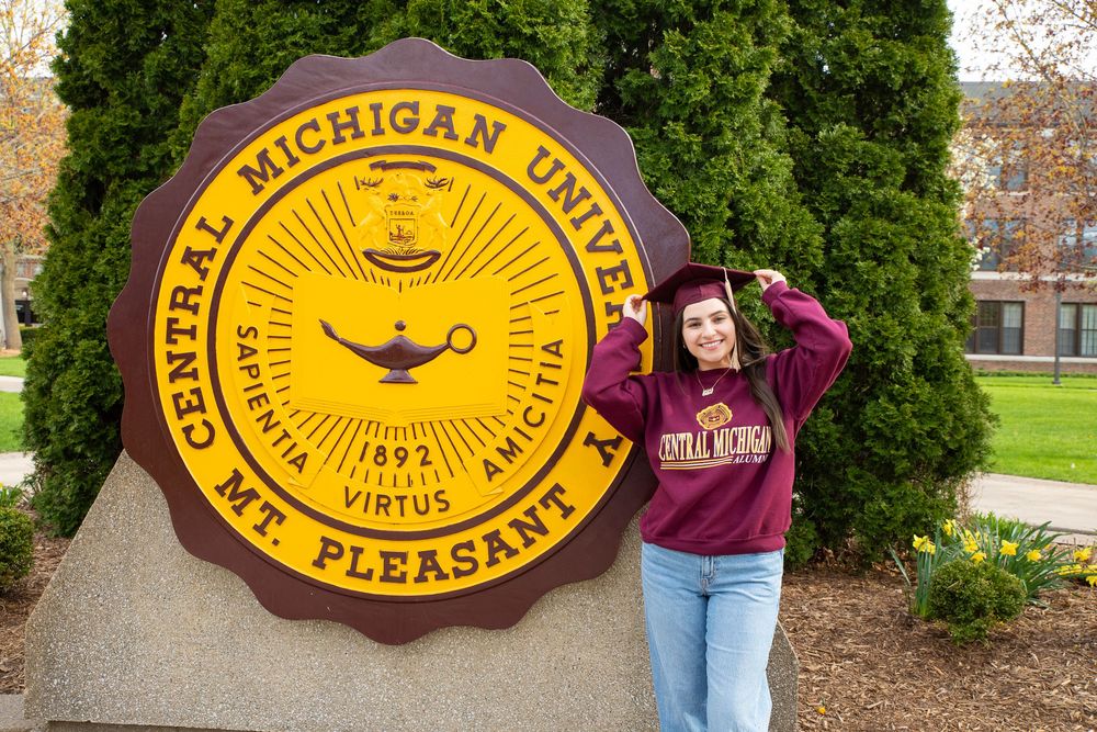 Graduate in cap and gown poses proudly beside Central Michigan University seal monument in Mt. Pleasant campus.