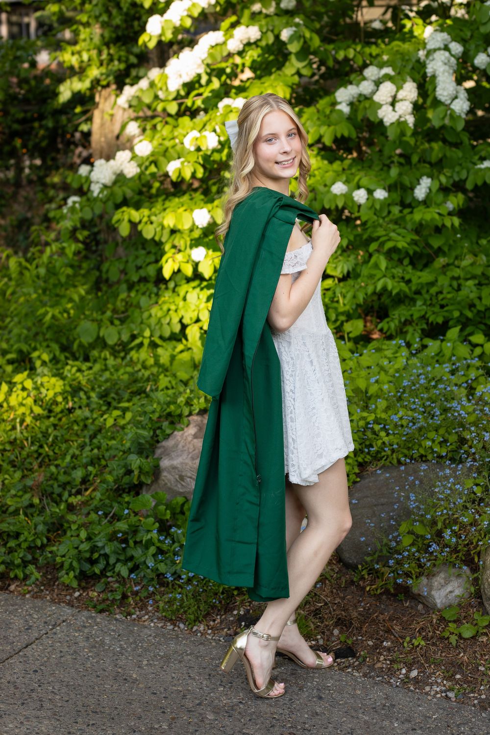 Young blonde woman in white dress holding green graduation gown, posing outdoors among lush green bushes and white flowers.