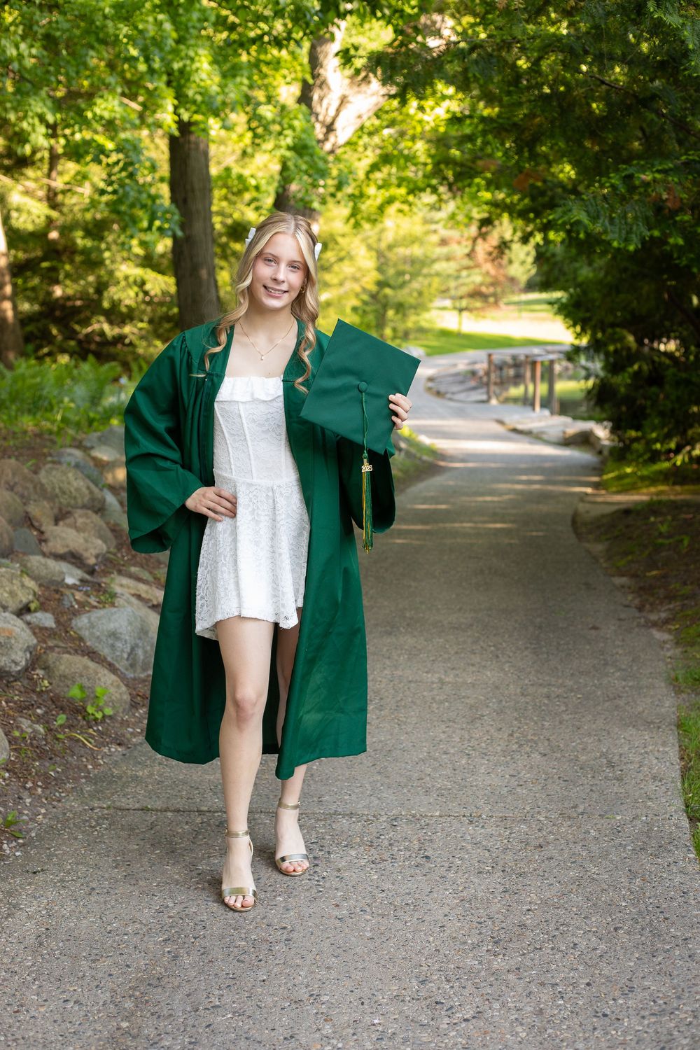 Smiling blonde graduate in green cap and gown over white dress poses on a tree-lined path.