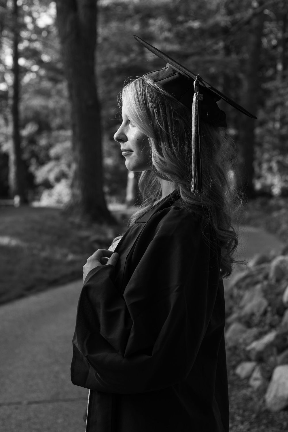 Black and white photo of a young woman in graduation cap and gown standing outdoors among trees.