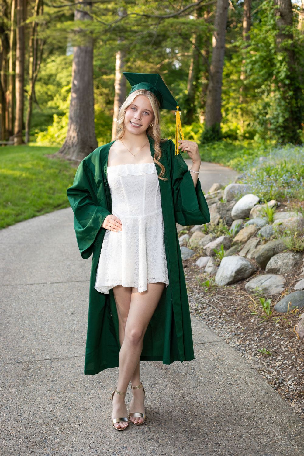 Smiling blonde graduate in green cap and gown with tassel, wearing white dress and heels, posing outdoors on a garden path.