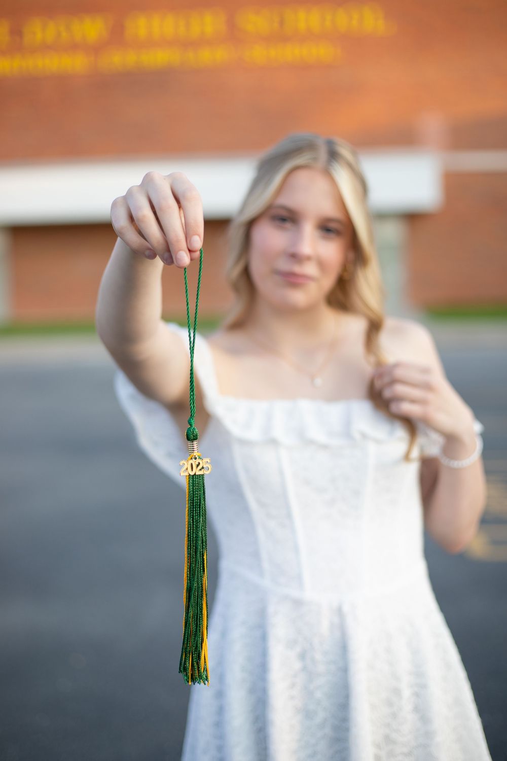 Blonde girl in white dress holding green and gold 2025 graduation tassel outdoors near school building.