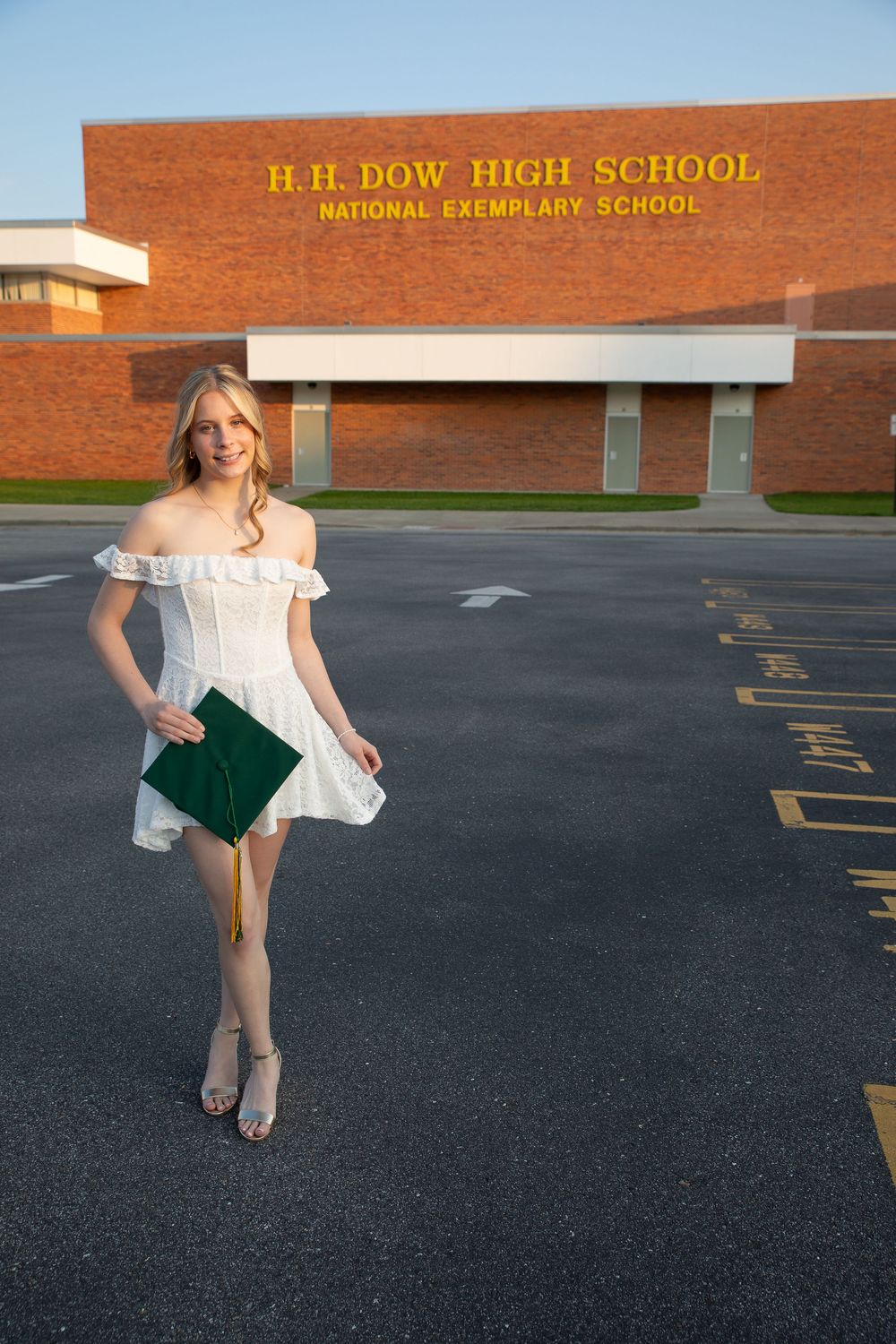 A young woman in a white dress holds a green graduation cap outside H.H. Dow High School on a sunny day.