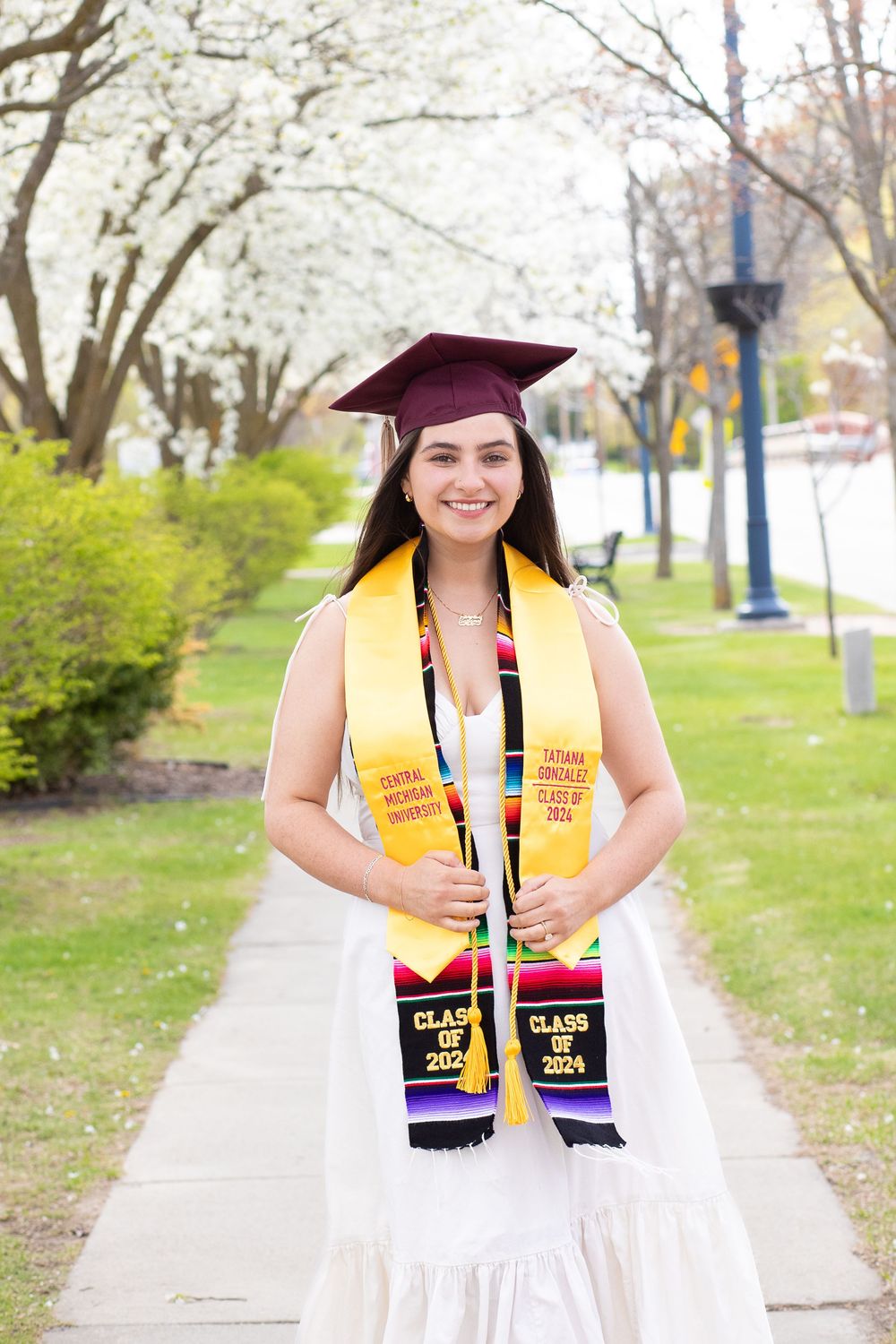 Smiling female graduate in maroon cap and white dress wearing Class of 2024 stoles outdoors among blooming trees.