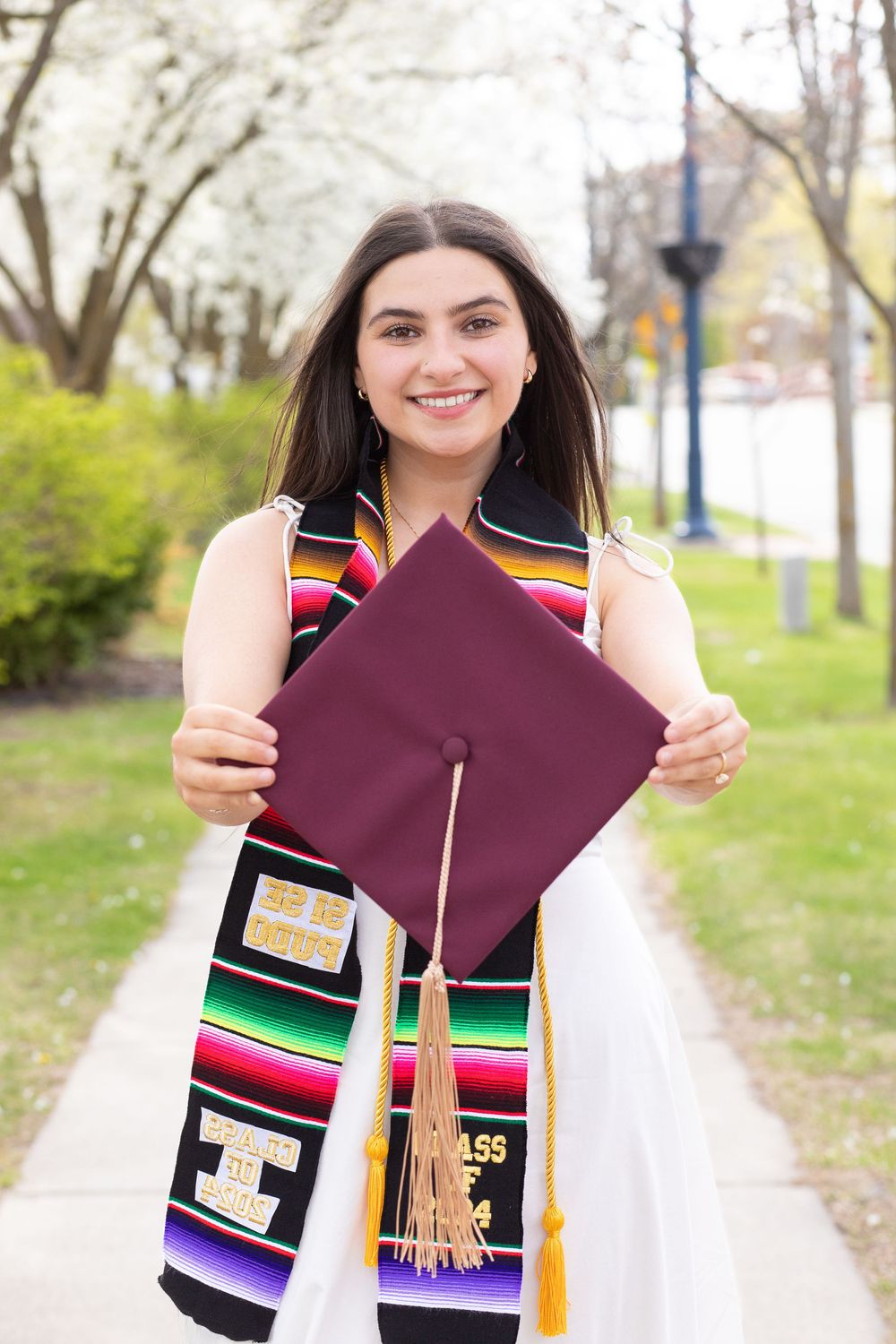 Smiling female graduate holding maroon cap with tassel, wearing colorful sash outdoors in spring.