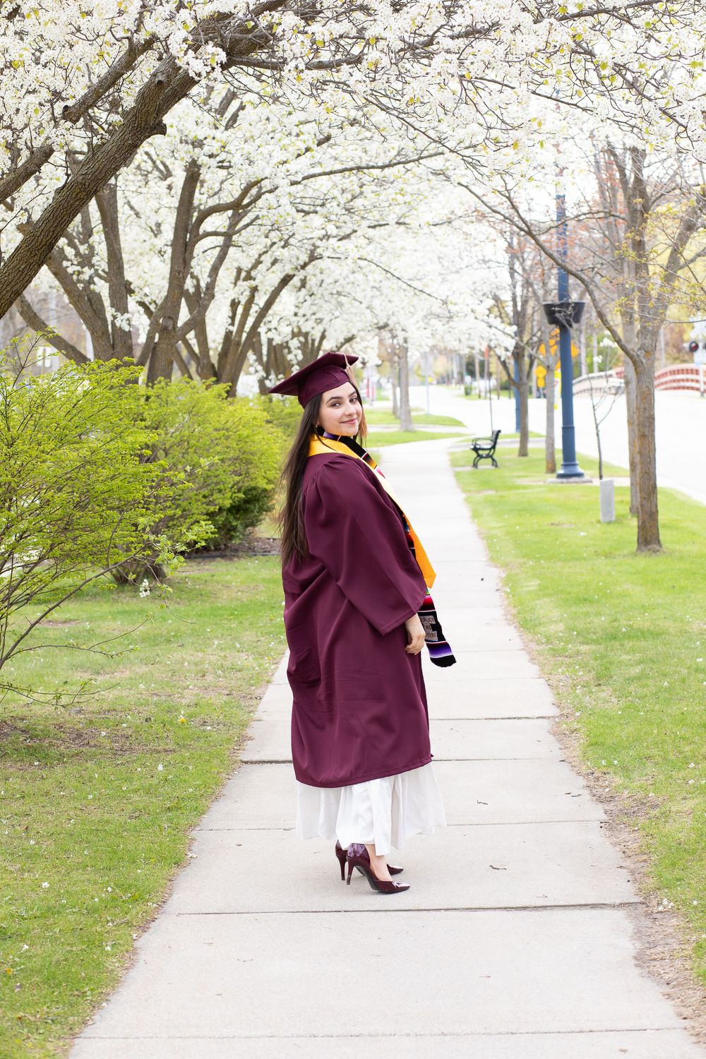Female graduate in maroon cap and gown poses on a tree-lined campus walkway with white blossoming trees in spring.