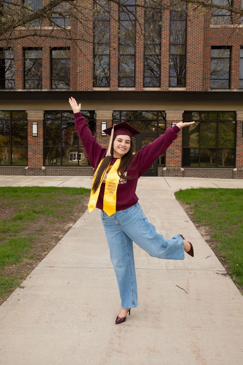 A joyful graduate poses in cap and gown with arms raised on a brick university campus walkway.